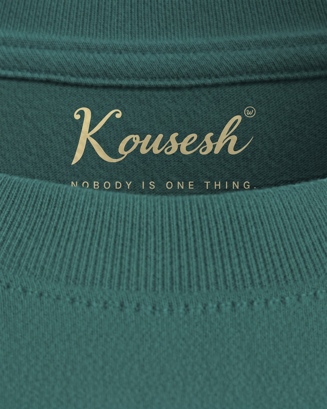 Inside the neckline, a pale yellow label printed into the cotton reading Kousesh and NOBODY IS ONE THING., partly hidden behind the front collar