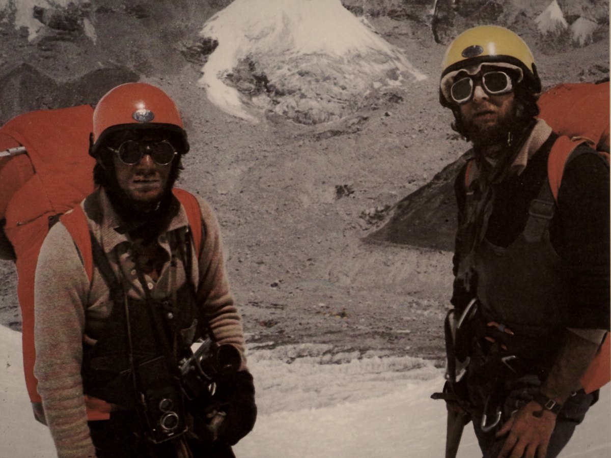 Two climbers in helmets and goggles standing on snow below a mountain face, 1980