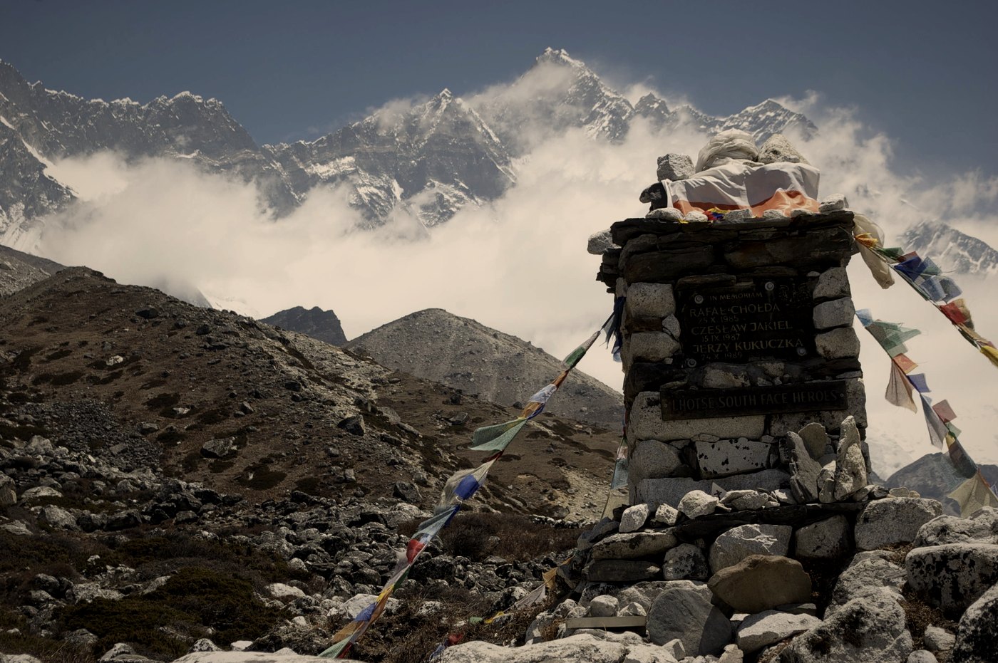 A stone memorial with prayer flags on a rocky slope, with a snowy mountain face rising behind it in cloud